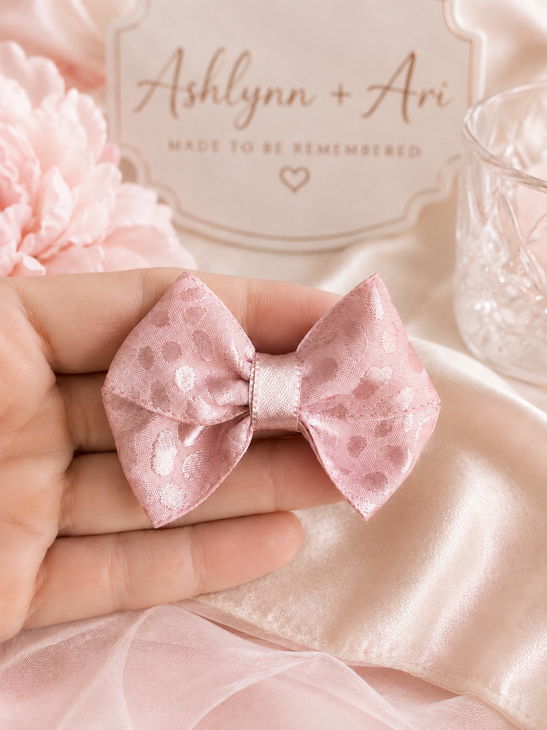 A small pink satin bow with a subtle polka dot pattern is held in a person's hand against a soft pink fabric backdrop with decorative flowers and a glass.
