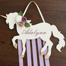 A personalized wooden unicorn wall hanging with a white body, pink and lavender stripes hanging below, faux flowers on the neck, and the name 'Adalynn' written in script. It is displayed on a dark wood background.