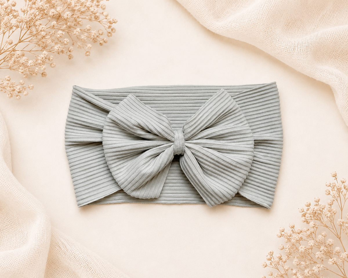 A ribbed fabric baby headband with a large bow, laid flat on a warm cream background with decorative dried flowers and soft draped fabric.