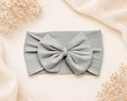 A ribbed fabric baby headband with a large bow, laid flat on a warm cream background with decorative dried flowers and soft draped fabric.