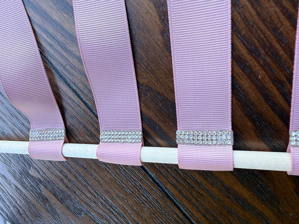 A close-up of a lavender pink grosgrain ribbon looped around a white dowel, decorated with a row of silver rhinestone embellishments, set against a dark wood background.