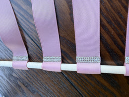 A close-up of a lavender pink grosgrain ribbon looped around a white dowel, decorated with a row of silver rhinestone embellishments, set against a dark wood background.