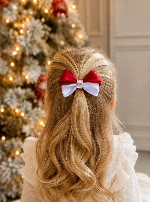 A young girl viewed from behind, wearing a red and white holiday bow in her blonde hair, with a decorated Christmas tree and warm lights in the background.