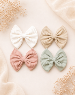 Four pleated fabric hair bows in white, beige, dusty pink, and sage green, arranged in a flat lay on a warm cream background with soft fabric and dried flowers.