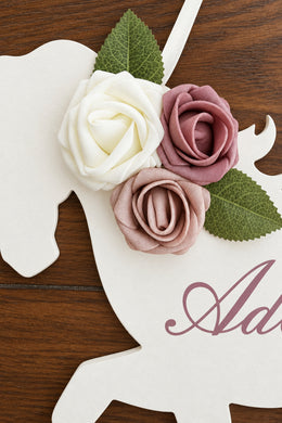 A close-up of a white wooden horse-shaped decoration or sign on a dark wood surface, adorned with three fabric roses in white, dusty pink, and mauve, with green leaves. Partial script lettering is visible on the sign.
