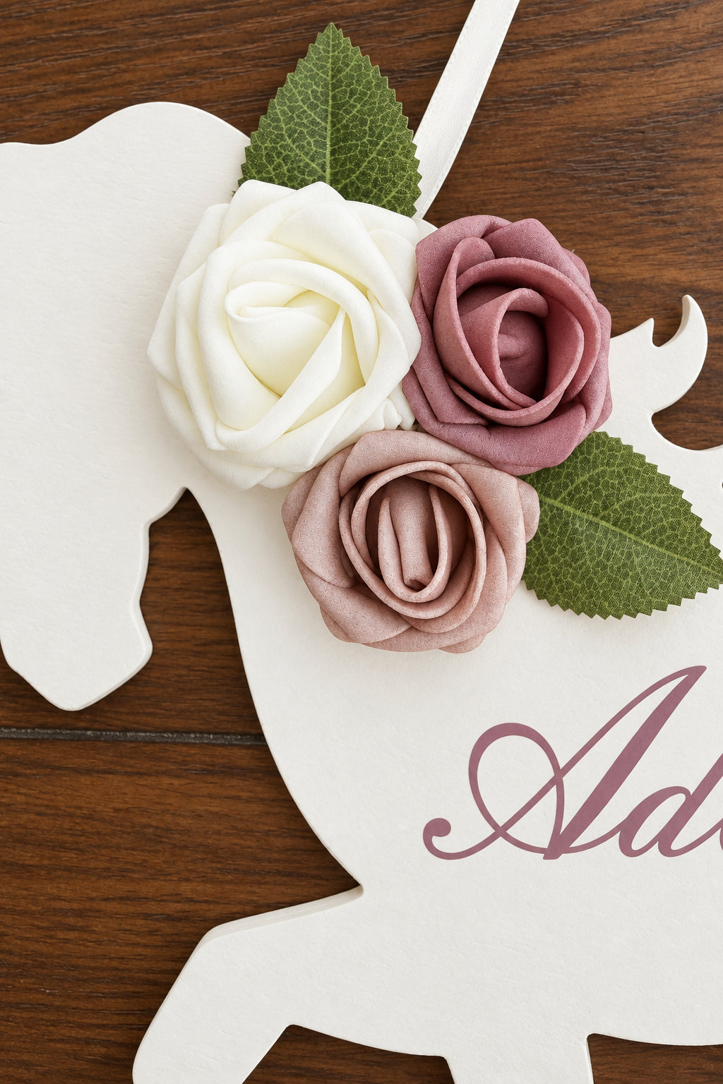 A close-up of a white wooden horse-shaped decoration or sign on a dark wood surface, adorned with three fabric roses in white, dusty pink, and mauve, with green leaves. Partial script lettering is visible on the sign.