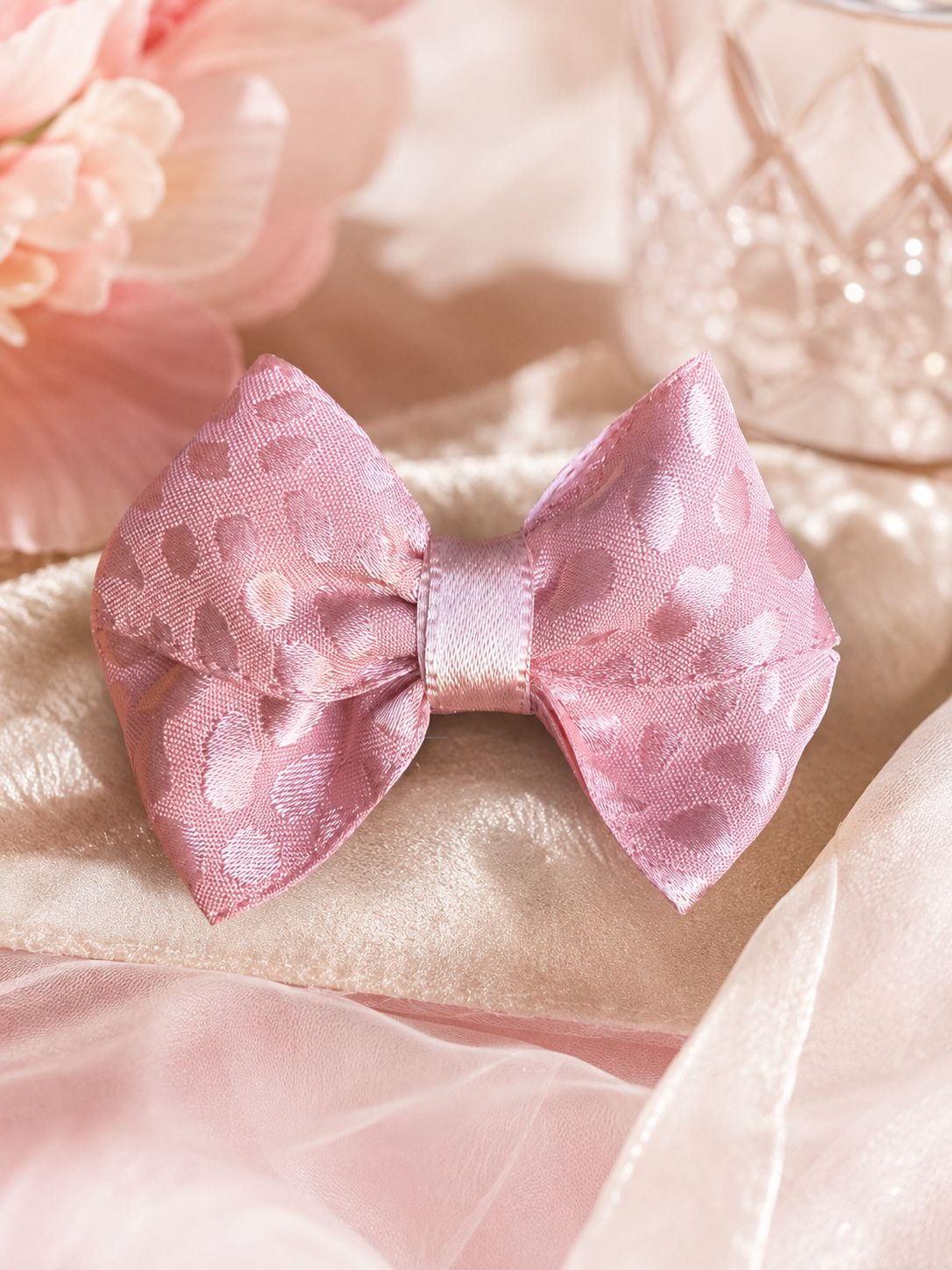 A pink satin bow with a subtle heart pattern, styled on a soft beige fabric with pink flowers and a glass in the background.