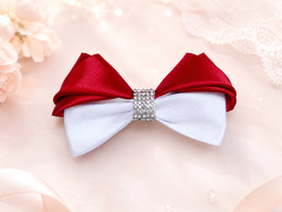 A satin bow hair clip with a red top half and a white bottom half, centered with a rhinestone-embellished band, displayed on a soft beige background with decorative lace and flowers.