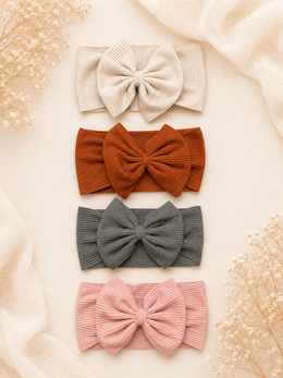 Four ribbed bow headbands in cream, rust orange, charcoal gray, and dusty pink, arranged vertically on a warm beige background with soft draped fabric and dried floral accents.