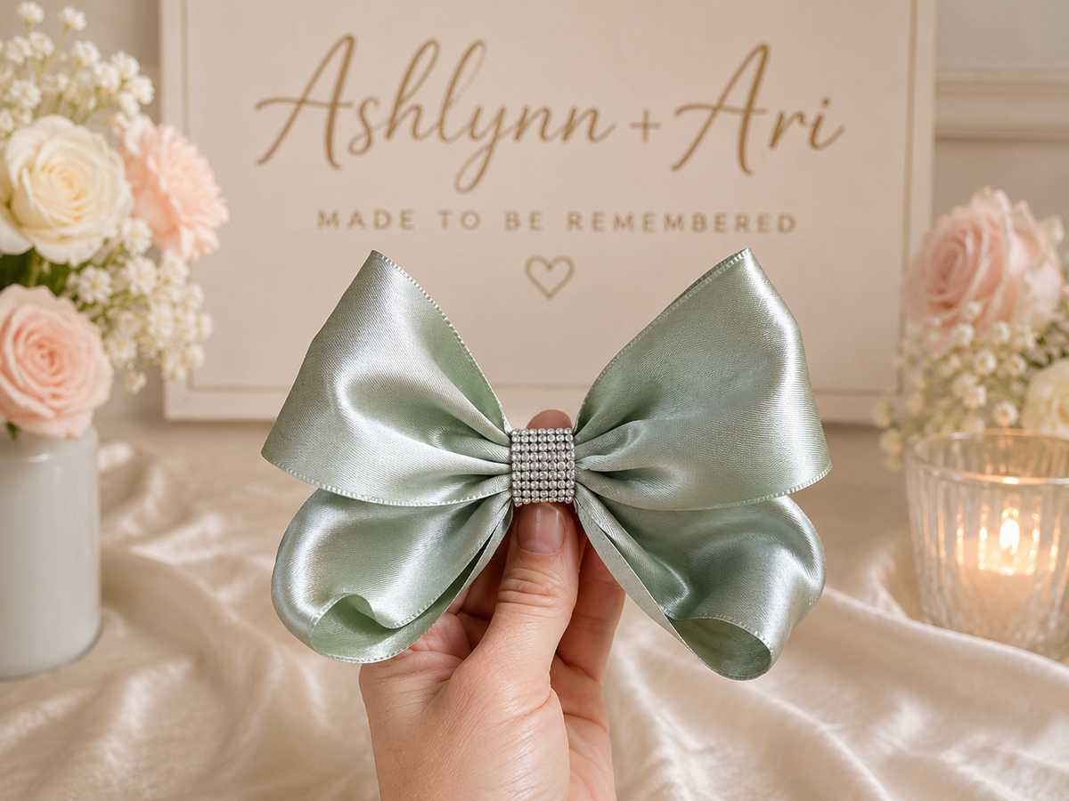 A sage green satin bow with a rhinestone-embellished center, held by a hand against a warm, decorated backdrop with flowers and a sign reading 'Ashlynn + Ari'.