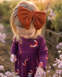 A toddler girl wearing a large rust-orange corduroy bow headband and a purple long-sleeve dress with fox prints, standing in a garden among small pink flowers.
