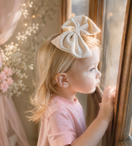 A young child with blonde hair wearing a cream and white striped fabric bow headband and a pink t-shirt, looking out a window.