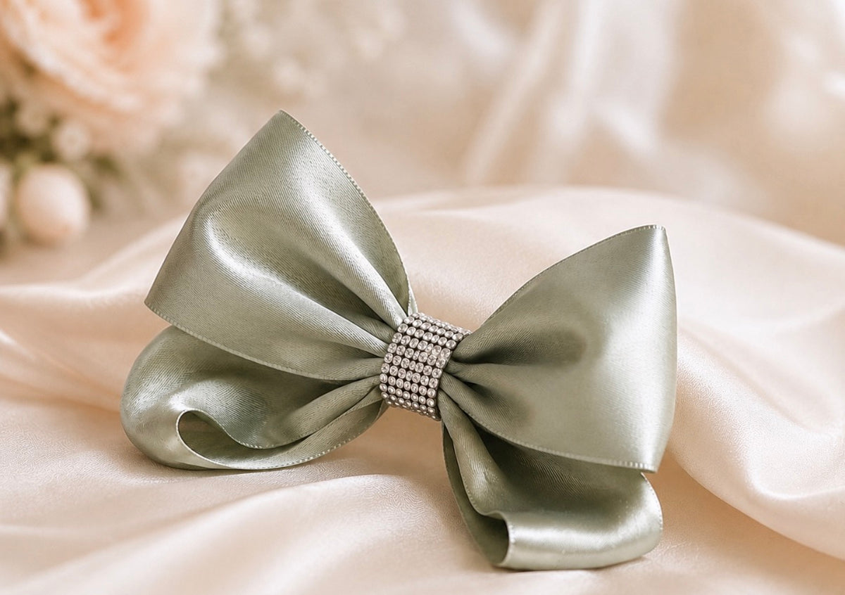 A large satin bow in a muted sage green color, featuring a decorative rhinestone-embellished center band, displayed on draped cream satin fabric.