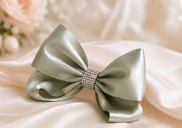 A large satin bow in a muted sage green color, featuring a decorative rhinestone-embellished center band, displayed on draped cream satin fabric.
