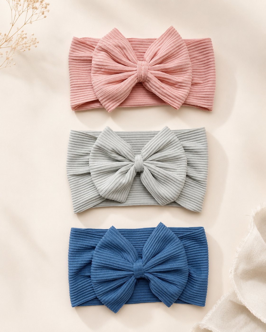 Three ribbed fabric bow headbands in dusty pink, light blue, and medium blue, arranged vertically on a warm cream background with dried floral accents.