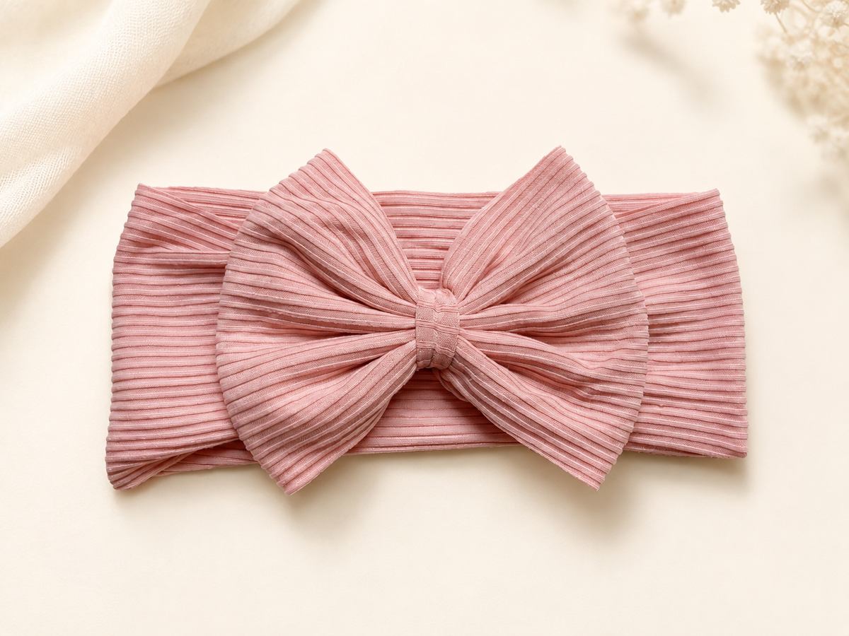 A dusty pink ribbed fabric headband with a large bow, laid flat on a light cream background with soft decorative fabric and dried flowers in the corners.
