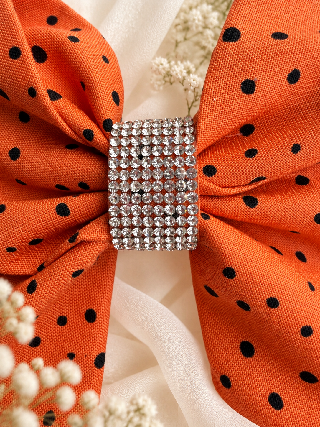 An orange fabric hair bow with black polka dots and a rhinestone-encrusted center, styled with white baby's breath flowers on a white fabric background.